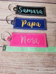 three personalized name tags are shown on a wooden surface, one is blue and the other is pink