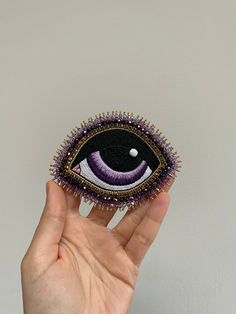 a hand is holding an embroidered brooch with purple eyes