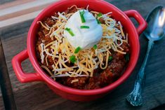 chili with sour cream and shredded cheese in a red bowl next to a spoon on a wooden table