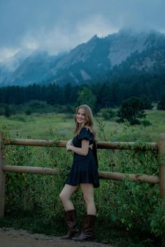 a woman standing next to a wooden fence in front of a mountain with trees and bushes