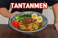 a person holding a bowl filled with noodles and meat on top of a wooden table