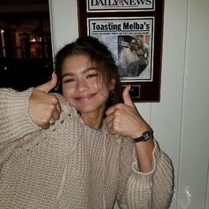 a woman giving the thumbs up sign in front of a news paper advertisement on a wall