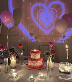 a table topped with a cake next to candles and balloons in front of a heart - shaped wall