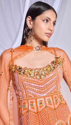 Our burnt orange 3-piece sharara set includes a peplum top, sharara, and dupatta. The peplum top features delicate embroidery with crystals, pearls, and sequins, complemented by gold metallic flowers on the front neckline and scalloped hem. The georgette sharara is adorned with crystal tassels, while the matching dupatta is embellished with scattered embroidery and finished with a delicate pearl and golden border. This ensemble offers a perfect blend of opulence and elegance. Designer Orange Palazzo Set With Zari Work, Orange Designer Palazzo Set For Navratri, Festive Designer Orange Sharara, Orange Palazzo Set With Dupatta And Traditional Drape, Festive Orange Designer Sharara, Traditional Drape Orange Palazzo Set With Dupatta, Designer Orange Sharara With Zari Work, Orange Bollywood Designer Palazzo Set, Orange Palazzo Set With Resham Embroidery