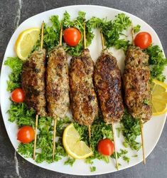 several skewered meats are on a plate with tomatoes, lettuce and lemon wedges