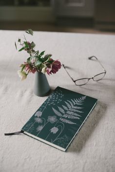 a book and flower vase sitting on a bed with eyeglasses next to it