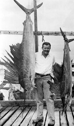 black and white photo of man holding two large fish on dock with fishing poles in background