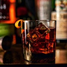 a close up of a glass on a table with liquor bottles in the background and an orange slice sticking out of it