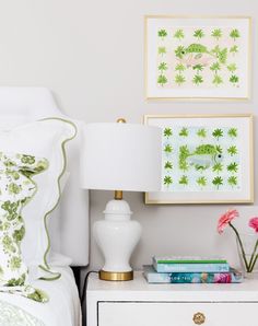 a white nightstand topped with books next to a lamp and pictures on the wall above it