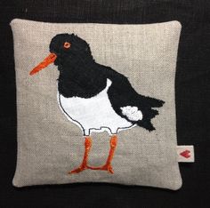 a black and white bird with orange legs on a gray pillow cover that is embroidered onto it