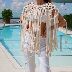 a woman standing next to a swimming pool wearing white pants and a crocheted shawl