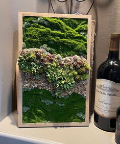 a bottle of wine sitting next to a painting on a wall with moss and flowers