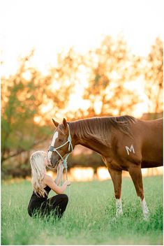 a woman kneeling down next to a brown horse on top of a lush green field