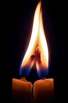 two lit candles in the dark with one burning and another being turned on by an orange flame