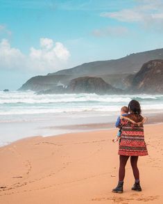 a woman standing on top of a sandy beach next to the ocean holding a baby