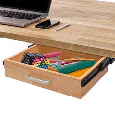 an open drawer underneath a desk with a laptop and pencils in it, on top of a wooden table