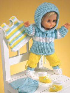 a doll is standing on a chair holding a knitted sweater and booties in front of her