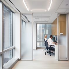 two people sitting at desks in an office with glass walls and large windows looking out onto the city