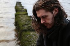 a man with long hair standing next to the water looking at his cell phone while wearing a black coat