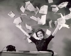 a woman sitting at a desk with her hands in the air and papers flying above her