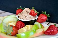 a plate topped with sliced apples, bananas and strawberries next to a bowl of chocolate frosting