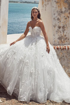 a woman in a white wedding dress sitting on a stone wall next to the ocean