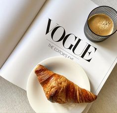 a croissant on a plate next to a cup of coffee and a magazine