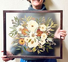 a woman holding up a framed painting with flowers on it's face and smiling at the camera