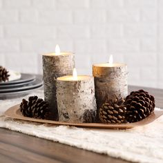 three lit candles sitting on top of a wooden tray next to pine cones and plates