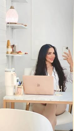 a woman sitting at a table with a laptop in front of her and holding a cell phone