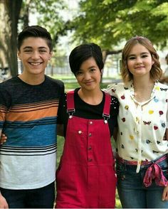 three young people standing next to each other in front of trees and grass with one person wearing overalls