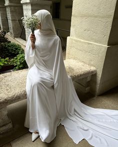 a woman dressed in white is sitting on a bench and holding a bouquet of flowers