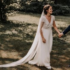 a bride and groom hold hands as they walk through the grass in front of trees