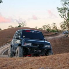 a black suv driving down a dirt road next to a tree and grass covered hill