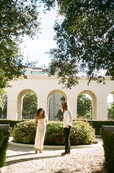 a man and woman standing next to each other in front of some hedges with trees