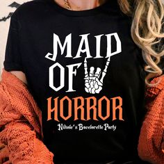 a woman wearing a black shirt with the word maid of horror printed on it and an orange sweater