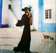 a woman in a long black dress and hat is walking with a dog on the street