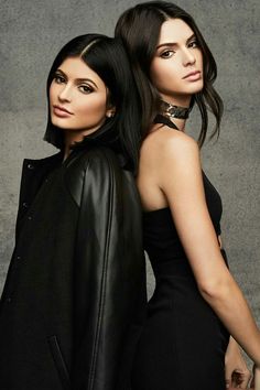 two beautiful women standing next to each other in front of a gray wall and one is wearing a black dress