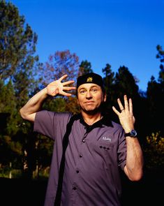 a man in purple shirt and black hat holding his hand up to the sky with trees behind him