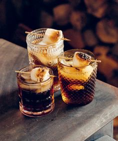 three glasses filled with different types of drinks on top of a wooden table next to firewood logs