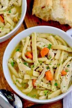 two bowls filled with chicken noodle soup on top of a wooden table next to bread