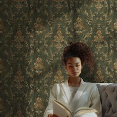 a woman sitting on a couch reading a book in front of a floral wallpaper