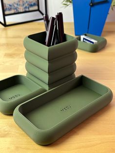 three green trays with pens and pencils in them on a wooden table next to a clock