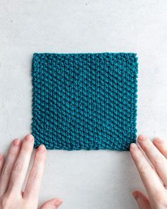 two hands holding up a knitted square on top of a white surface with blue yarn