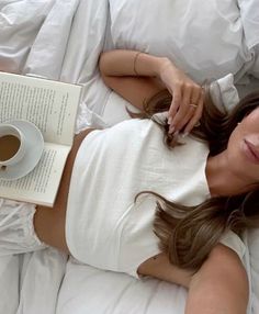 a woman laying in bed with an open book and cup of coffee