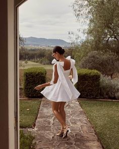 a woman in a short white dress is walking down a path with her back to the camera