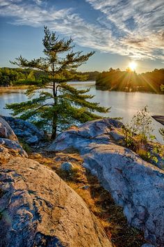 the sun is setting over a body of water with rocks and trees in front of it