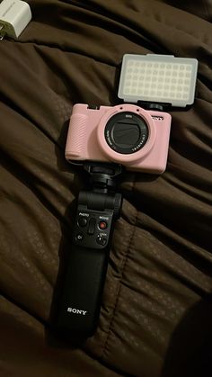 a pink camera sitting on top of a bed next to a flash light and remote control