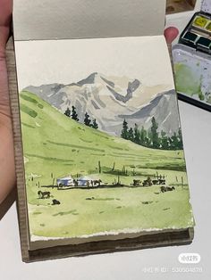 a person holding up a piece of paper with watercolors on it and mountains in the background