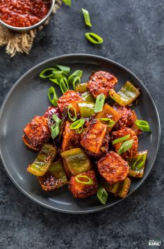 a black plate topped with fried vegetables next to a bowl of chili sauce and green onions